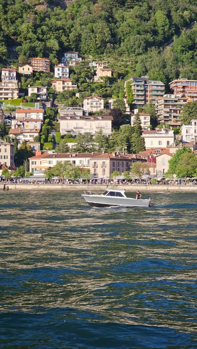 The image shows an aerial view of a shoreline in Como, Italy. A mix of residential buildings and hotels with orange, tan and white exteriors line the shore, interspersed with trees and manicured green spaces. A waterfront promenade runs along the edge of the buildings. People are sparsely positioned along the promenade. In the water a modern boat with large rectangular windows, travels parallel to the shore, creating a light white wake on the blue water. The reflections of the buildings are visible on the water's surface. A steep, densely forested hillside rises on the opposite side of the buildings.