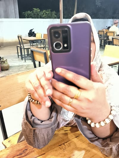 The image depicts a person, presumably female, holding a purple smartphone in front of them, likely taking a photo or video. The person is wearing a hijab, a gray patterned blouse, and bracelets on both wrists. Gold rings are visible on one hand. The person is seated at a wooden table in what appears to be an outdoor dining area. In the background, other tables and chairs are visible, along with a low wall and some foliage. Further in the background, a man wearing a red cap is seated. A gas tank sits to the left, near the edge of the photo. The location may be Cempeudak, Indonesia.