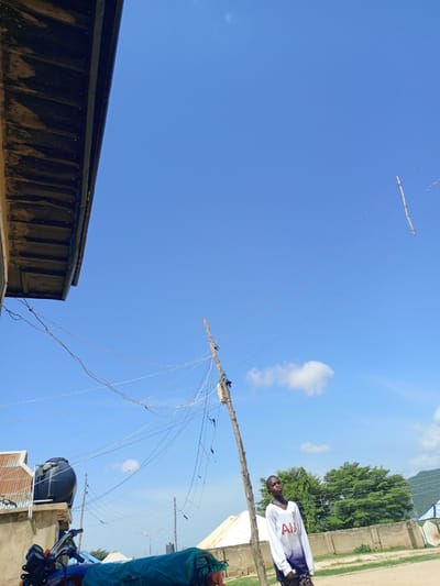 In Barakin Babele, Nigeria, under a clear blue sky with a few wispy clouds, a young boy stands near a wooden utility pole adorned with electrical wires. He is wearing a white long-sleeved shirt with red and blue accents and patterned dark pants. To the left of the frame, the underside of a dark, weathered roof overhang is visible. In the background, a large black water tank sits atop a concrete structure, with a motorcycle parked in front of it, partially covered by a green tarp. A concrete wall runs across the middle ground, with lush green trees and hills in the distance. Another utility pole, also covered in wires, is situated closer to the center of the image. The scene appears to be an outdoor, possibly residential area.