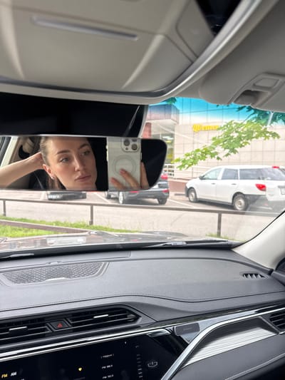 A young woman is reflected in the rearview mirror of a car. She is looking at her phone, which she is holding in her right hand. Her left hand is resting on the back of her head. She is wearing earrings and a black shirt. The car's interior is visible in the foreground, with a dashboard and steering wheel. Outside the car, a white station wagon is parked. The scene appears to be a parking lot in front of a building with a sign that says "Цветы" (Flowers) in yellow letters. The location is the city of Korolyov, Russia. The weather appears to be overcast.