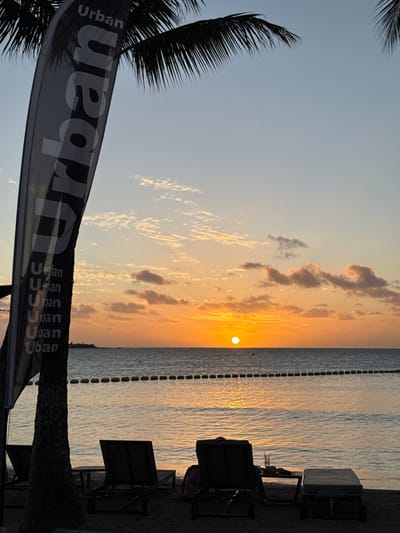 The image depicts a sunset over a coastal beach in Noumea, France. The sun, a bright orange circle, is positioned low on the horizon above the calm ocean, reflecting orange and yellow light across the water and into the partly cloudy sky. A line of yellow spherical buoys is visible horizontally across the water. In the foreground, silhouetted elements include a tall, dark vertical flag featuring the word "Urban" repeated multiple times along its length, standing beside a palm tree trunk on the left. To the right, two dark-toned beach lounge chairs face the ocean. Adjacent to the rightmost chair are a small dark table, holding a transparent glass, and a dark cooler. No individuals are visible.
