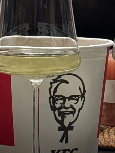 A close-up shot focuses on a partly-filled, clear champagne flute positioned in front of a KFC (Kentucky Fried Chicken) bucket. The iconic Colonel Sanders logo is prominently displayed on the bucket. The flute is angled, obscuring a portion of the logo. The background includes hints of other objects, including a wicker basket and a dark surface. The setting appears to be indoors. No people or other subjects are visible. The location, Zovuni, Armenia, cannot be confirmed through this image.