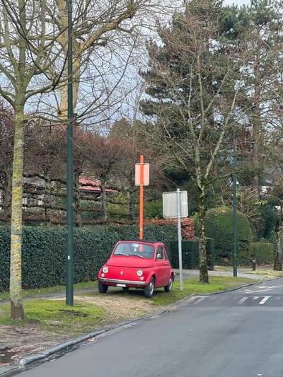 This image depicts a static outdoor scene in a suburban or residential area within Uccle - Ukkel, Belgium, on an overcast day, likely during late autumn or winter. The primary subject is a vintage red Fiat 500 automobile, facing slightly left and angled towards a paved road. The vehicle is parked on a grassy and gravel shoulder adjacent to the road.
The paved road, dark grey in color, runs across the foreground, separated from the shoulder by a concrete curb. Road markings, possibly indicating a speed bump, are visible on the road surface to the right. On the shoulder, a tall, dark green utility pole stands near the left edge of the frame. Further along the shoulder, behind the red car, an orange pole supports two vertically aligned, rectangular, light-colored signs. To its right, a grey metallic pole holds a single, rectangular, light-grey sign.
Behind these elements, a dense, dark green hedge delineates a property line. Above the hedge and extending into the background, numerous deciduous trees with bare branches are visible, interspersed with some evergreen foliage, including a large coniferous tree on the right. Portions of residential structures with red-tiled roofs are faintly visible through the bare trees in the distant background. No individuals or animals are present in the scene.