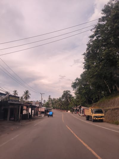 The image captures a slightly elevated perspective of a curved asphalt road in a rural or semi-urban setting in Seulimeum, Indonesia, under an overcast sky displaying soft pink and grey hues. Power lines stretch horizontally across the upper part of the frame.

On the left side of the road, a series of simple, single-story buildings with corrugated metal roofs and open storefronts line the street. Palm trees and other tropical foliage are visible behind and between these structures. A person is seated in one of the open storefronts. Further down the road on the left, a blue motorized pedicab (becak) with a driver is in motion.

The right side of the road features a steep, densely tree-covered hillside, with a stone retaining wall at its base. A bright yellow Mitsubishi FUSO dump truck is parked on the road shoulder next to this wall. The brand "MITSUBISHI" is visible on the truck's front grille. The dense trees on the hillside are lush and green.

The soft, diffuse lighting and the sky's coloring suggest either early morning or late afternoon, or a heavily cloudy day. The scene depicts a quiet local street with typical roadside establishments and tropical vegetation.