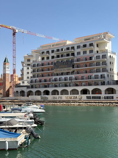 Here is a detailed description of the image:
**Scene Overview:**
The image shows a waterfront scene in Eilat, Israel, dominated by a large, modern building with multiple balconies and windows. The building is labelled "VENEZIA YOSSI AVRAHAMI & ALMOGIM". This likely indicates a residential or commercial complex. In the foreground, several boats are docked along the waterfront. A red and white construction crane is visible in the background, suggesting ongoing construction or development.
**Objects and Infrastructure:**
- Large, white building with multiple floors, balconies, and windows.
- Waterfront with a rocky barrier.
- Several docked boats of varying sizes and designs.
- Red and white construction crane.
- A tower with a green roof and an orange structure below, possibly a replica or themed architecture.
**Environment:**
The scene is bathed in daylight, suggesting a clear, sunny day. The water appears calm and green.
**Location:**
The presence of the "VENEZIA YOSSI AVRAHAMI & ALMOGIM" building and other visual cues suggests this is likely located in the city of Eilat, Israel, near the waterfront or marina area.