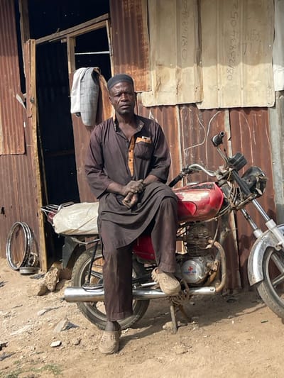 An adult male is seated astride a red "CARGO" brand motorcycle in an outdoor setting in Chikaji, Nigeria. The man, with a dark complexion, wears a dark brown long-sleeved tunic with a lighter brown chest panel and a dark skullcap. He has a short beard and mustache, and his hands are clasped together, appearing soiled. He wears light brown Crocs-style clogs. The motorcycle is visibly used, featuring a red fuel tank, chrome exhaust, and a white, cloth-wrapped object secured to its rear rack; its kickstand is deployed. Behind the man and motorcycle is a structure composed of rusty, corrugated metal sheets, indicating wear. An open doorway, framed by wooden planks, is visible to the man's left, with a light-colored, patterned jacket hanging on the wall nearby. Vertical numbers, including "0813083405" and "08/20834052", are inscribed in white on the corrugated sheets to the man's right. The ground is uneven, dusty dirt, and two chrome motorcycle wheel rims lean against the corrugated wall to the left of the motorcycle.