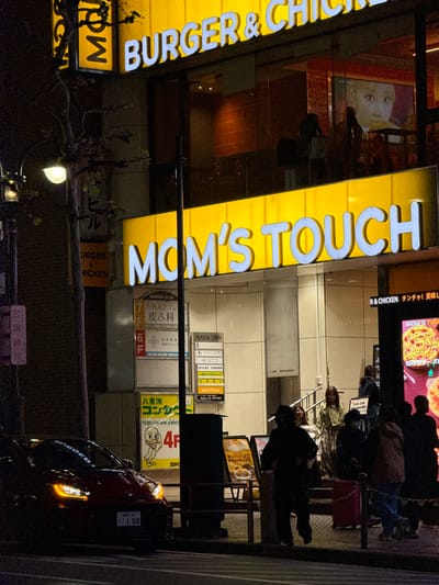 An urban street scene unfolds at night, likely in Shibuya, Japan, characterized by a brightly illuminated building housing various businesses. The central focus is a multi-story commercial building with a prominent yellow sign displaying "MOM'S TOUCH" and "BURGER & CHICKEN" in large, glowing letters. Reflections of people and interior lighting are visible in the building's glass facade.
Several people are gathered on the sidewalk in front of the building, some appearing to be queuing or waiting. Their forms are largely silhouetted against the bright signs and interior lights, making individual features indistinct.
Below the main fast-food signs, smaller signs in Japanese advertise other tenants of the "MARUDAI BLD." These include "渋谷スクラン 皮ふ科" (Shibuya Scramble Dermatology) on the 7th floor, "KARA 内科クリニック" (Kara Internal Medicine Clinic) on the 6th floor, a pharmacy ("処方せん受付 渋谷駅前薬局"), and "八重洲コンタクト" (Yaesu Contact) with a cartoon duck and web address "http://www.yaeco" on the 4th floor. Other text mentions "R & CHICKEN チンチャ! 美味し" (Really! Delicious!) and "MOM'S TOUCH PLUG IN STUDIO" alongside images of food like pizza. A "NEW! ・チキンスティック 450" (Chicken Sticks 450) offer is visible, as well as a sign indicating "21:30-22:00 Take away ONLY."
A dark-colored car, possibly red, is parked or driving on the street in the foreground to the right, with its headlights and taillights on. Its Japanese license plate is partially visible, reading "*練馬347* ・1 08". A bare-branched tree with some foliage and a streetlight illuminate the lower left corner. The overall ambiance is busy and energetic, typical of a city's nightlife.
