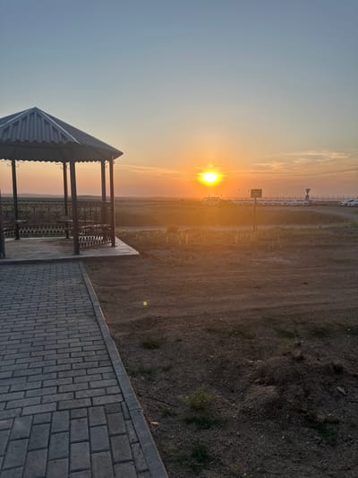 The image captures a sunset over the city of Bozshakol, Kazakhstan. In the foreground, a paved walkway made of gray bricks leads to a hexagonal gazebo with a corrugated metal roof supported by dark metal posts. The gazebo features decorative metal railings and benches inside. The ground around the gazebo is bare earth with some sparse vegetation.
The middle ground reveals a fenced-off area with a dirt road. Beyond the fence, a row of white vehicles, possibly cars or vans, is parked. The setting sun, a bright orange disc, is positioned low in the sky, casting a warm glow and long shadows across the landscape. The sky transitions from a pale blue at the top to shades of orange and yellow near the horizon, with a few wispy clouds. The overall environment appears to be an open, somewhat arid landscape.