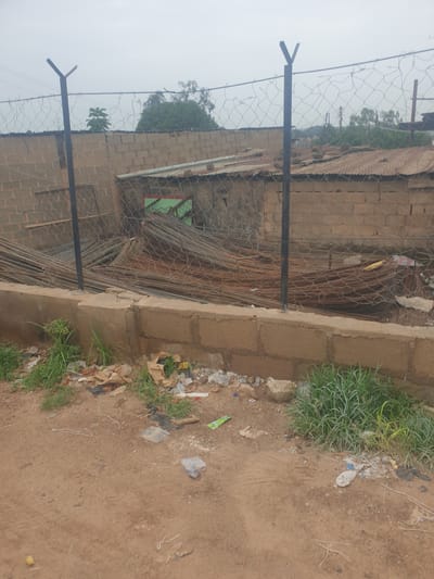 This image depicts an outdoor scene with a fence in the foreground, made of metal posts and wire mesh. Beyond the fence, there is a brick wall and a corrugated metal roof. A large pile of metal rods is visible behind the fence. The ground in front of the fence is dirt and littered with various debris, including plastic bags, paper, and plant matter. Some grass is growing on the side. The sky is overcast. The scene appears to be an undeveloped or industrial area. There is no indication this image is located in Washington, United States of America; it is highly unlikely to be from Washington D.C. or Washington State given the visual context.