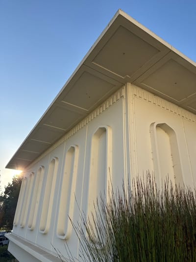 A low-angle, exterior shot captures the corner of a modern, white building under a clear blue sky. The building features a distinctive overhang with recessed square panels on its underside. The facade is adorned with a series of tall, narrow, arched recesses, evenly spaced along the side. The sun is setting, casting a lens flare through the trees in the background and illuminating the side of the building. In the foreground, tall, thin, dark green grass with reddish-brown tips grows in front of the building. A few parked cars are visible in the distance through the trees. The image appears to be taken in Glendale, United States of America.