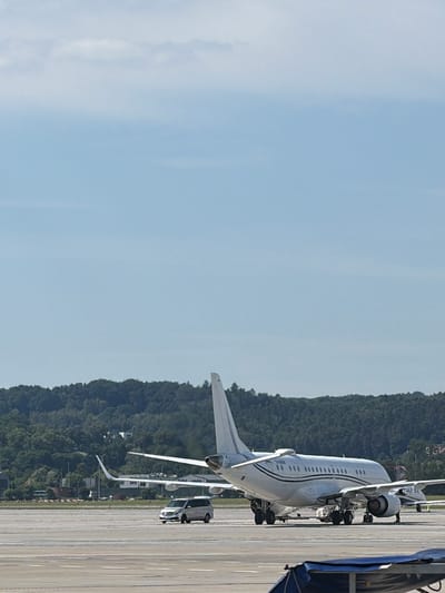 The image shows a private jet on an airport tarmac, with a lush, green hillside in the background. The jet is white with a black stripe running along its fuselage and appears to be parked. A white car is visible near the rear of the jet, and a section of an airport structure, possibly a terminal or a maintenance shed, is on the right side of the image. The sky is a clear, light blue, suggesting a bright, sunny day. The sun appears to be high in the sky, indicating it is likely midday. The location cues, such as the tarmac and the presence of an aircraft, confirm it is an airport. The overall scene is calm and professional, typical of an airport environment. There is no visible text in the image.