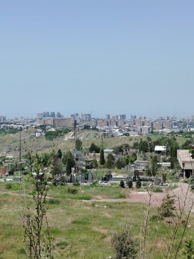 The image shows a daytime view of a cemetery in Jrvej, Armenia, with a city skyline in the background. The cemetery is filled with gravestones and mausoleums, some of which appear to be made of stone and metal. The ground is covered in grass and dirt paths. In the distance, there is a sprawling urban landscape with numerous multi-story buildings, including residential apartments and what appears to be construction sites. A large electrical pylon stands on a hill between the cemetery and the city. The sky is clear and blue. The overall scene suggests a juxtaposition of a place of remembrance with the ongoing life of a modern city.