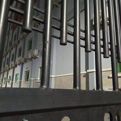 The image shows a building with multiple windows and air conditioning units, viewed through a metal fence. The building appears to be several stories high and is painted in a light blue and beige color scheme. The windows are rectangular and have green frames. The air conditioning units are white and are mounted below the windows. The metal fence is made of vertical bars and horizontal supports. The fence is black and appears to be made of metal. The scene is set at night or in low light conditions, as indicated by the shadows and the artificial lighting. The location of the image is the city of Ibadan, Nigeria.