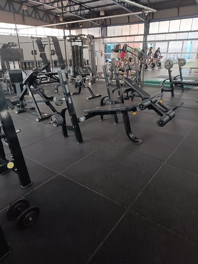 A gym interior showcases various weightlifting machines and equipment. The foreground features a leg extension machine and a bench press with weights. In the background, several individuals are visible riding stationary bikes near a window. The gym floor is covered with dark rubber tiles. The building appears to have a high ceiling with exposed metal beams and skylights, creating a bright and airy atmosphere.