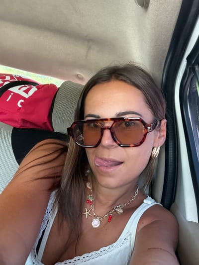 A woman in sunglasses and a white top poses inside a vehicle. She wears a gold charm necklace with a variety of pendants. A red bag rests on the car seat behind her. The interior of the vehicle is visible, including the ceiling, window trim, and headrest. The woman has brown hair and is wearing gold earrings. The light suggests it is daytime.