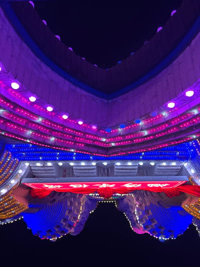 Captured from a ground-level perspective at night, the image showcases a brightly illuminated gate structure in Baqiao, China. The structure features a prominent arch on the left, outlined with purple lights, contrasting with rows of pink and blue lighting strips along its facade. A vertically oriented red sign with white Chinese characters is positioned towards the right, adorned with red decorative elements. The gate structure on the right side has a layered appearance, illuminated with blue and yellow lights. The background is a dark night sky, accentuating the vibrant colors of the gate's illumination.