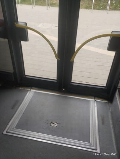 This image shows the interior of a public transport vehicle, likely a bus or tram, in Gdynia, Poland. The scene is viewed from inside, looking out through a large window and towards the entrance area.

The main subjects are the interior elements of the vehicle. Prominently featured are the window frames, a yellow grab handle, and the floor area around the entrance. The floor has a textured, dark grey mat with a decorative, silver-colored trim that forms a rectangular border. In the center of the mat, there appears to be a small, recessed circular object, possibly a latch or a cover. The window is large and appears to be tinted, showing a blurred view of the exterior. Outside, a paved surface with some grass is visible, suggesting the vehicle is stopped at a station or on a street.

The activity taking place is simply the static representation of the vehicle's interior, implying it is either stopped or stationary. There is no visible human activity.

Notable details include the time stamp in the upper right corner, which reads "29 черв. 2026 р., 17:06". This translates to "June 29, 2026, 5:06 PM", indicating the image was taken on a late afternoon in the summer. The lighting suggests it is daytime. The condition of the floor mat shows some wear, with minor cracks visible around the central circular object. The yellow grab handle is curved and positioned to assist passengers.

The visible text is the date and time stamp in Cyrillic script. The location context is Gdynia, Poland, which is inferred from the provided information. The overall impression is one of functional public transport infrastructure.