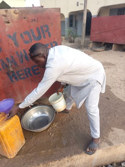 A man in a light-colored outfit turns a water tap on a rusty red water tank with the words "YOUR CLEAN WATER HERE." He holds a bucket and attempts to fill it with water, with an aluminum bowl and a yellow container nearby. The ground appears muddy. A building with an arched entrance is visible in the background.