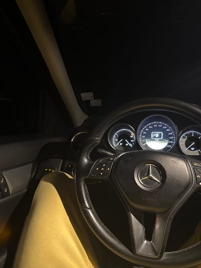The image displays the interior of a vehicle from the driver's perspective during low-light conditions, likely nighttime. A prominent black, leather-wrapped Mercedes-Benz steering wheel with a central three-pointed star logo and "AIRBAG" inscription is visible. The steering wheel features multi-function controls on both sides. Behind it, the illuminated instrument cluster shows a digital readout of "0 KM/H," indicating the vehicle is stationary, alongside an illuminated fuel gauge displaying a partial fuel level and an RPM gauge at zero. The lower part of a person's left leg, clad in light yellow or mustard-colored casual pants, is visible to the left of the steering wheel. Portions of the dark gray dashboard, a dark air vent, and the driver's door panel with control buttons are discernible. A light-colored A-pillar runs diagonally on the left, and two small, rectangular, light-colored objects are affixed to the dark headliner. No external features identifying the city of Vranentsi, Bulgaria, or any other specific geographical location are discernible within the image frame.