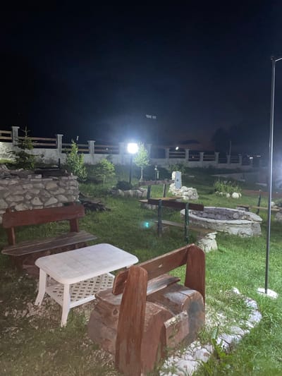 This image depicts an outdoor seating area at night, potentially a park or a rest stop, located in Javorje, Montenegro. The scene is illuminated by artificial lights, creating a somber ambiance. In the foreground, a white rectangular table with a patterned top is positioned on the grass. To the left of the table is a wooden bench. To the right, a unique bench constructed from logs and wood planks is visible.

Further back, a stone-lined fire pit with surrounding benches indicates a communal gathering spot. Various young trees and shrubs are scattered throughout the grassy area. A stone wall and a wooden fence form a boundary in the background, separating the seating area from what appears to be a road or another outdoor space. A lamppost stands to the right, providing additional light. In the distance, a dimly lit sign with text is visible. The overall environment suggests a quiet, rustic setting designed for outdoor leisure.