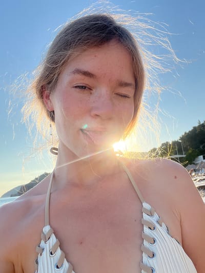 In Miljevci, Montenegro, a young woman is taking a selfie on a beach. She is wearing a white and beige bikini, and her hair is light brown and windswept. The sun is shining brightly, creating a lens flare that partially obscures her face. She is winking and sticking out her tongue, with the ocean and a hillside covered in trees in the background. There are beach chairs and umbrellas visible in the distance.