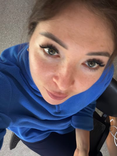 The image is a close-up, high-angle selfie of a young woman in Япрыково, Russia. She is looking directly at the camera with a neutral expression. The woman has fair skin with freckles, green eyes, dark eyebrows, and dark hair. Her eyelashes appear to be extended. She is wearing a bright blue fleece hoodie with a zipper, and dark pants. A portion of a black chair and some white wires are visible in the background. The lighting suggests it is daytime. The overall setting appears to be indoors, possibly an office or a home.