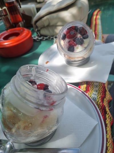 In Arbanassi, Bulgaria, on a table covered by a green cloth, two jars of dessert are displayed. The jar in the foreground, made of clear glass, contains a layered dessert with visible cake pieces and a creamy white filling, topped with raspberries and blueberries. To the left of this jar, a red ashtray is placed. In the background, another clear glass jar, similar to the first, holds a dessert topped with berries. A beige fabric object, possibly a purse, rests behind the jars. A woven textile with red, yellow, and black patterns is visible on the right side of the image. A spoon is positioned in front of the foreground dessert jar.