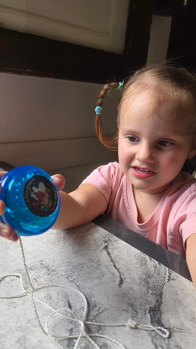 A young girl with light brown hair in two pigtails, secured with a blue and a green hair tie, is shown from the chest up, looking to her right with a slight smile. She is wearing a pink t-shirt and leaning on a grey and white speckled surface, possibly a table or counter. In her left hand, she holds a translucent blue yo-yo with a label featuring a cartoon character, possibly Mickey Mouse, and the words "SUPER YOYO". The yo-yo is connected to a white string that is tangled on the surface in front of her. The background is dimly lit, with a dark wooden structure above and a lighter, textured wall or surface behind the girl. The scene appears to be indoors. This image does not contain sufficient information to determine its location in Guía de Isora, Spain.