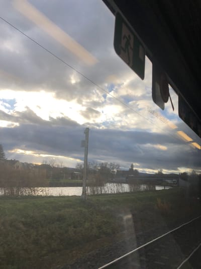 The image captures an exterior view from the interior of a train, looking out over the landscape of Dettwiller, France. In the upper right quadrant, a portion of the train's interior is visible, including reflections on the window pane. These reflections clearly show a green and white emergency exit sign depicting a running human figure, an orange emergency pull handle, and linear reflections of interior lighting.
Below the window, the train tracks are visible in the lower right, comprising ballast and metal rails. Immediately adjacent to the tracks, a strip of green grass and low-lying vegetation extends towards a body of water, likely a canal or river, which occupies the central left portion of the image. The water surface reflects a bright, elongated strip of sunlight, contrasting with the darker reflections of the sky and surrounding foliage. A tall, light-colored utility pole stands upright between the track and the waterway.
On the far bank of the water, several buildings with pitched roofs, indicative of residential or rural structures, are situated amidst autumn-toned trees and sparse vegetation. Further down the waterway, a multi-arched bridge crosses the water, and several white vehicles, possibly vans or trailers, are parked nearby. Distant hills are vaguely discernible on the horizon.
The sky is characterized by an expanse of dramatic, heavily overcast clouds, with patches of bright sunlight breaking through and illuminating the upper left quadrant. The overall lighting suggests either early morning or late afternoon. No individuals, specific actions, or interactions are observed within the frame.