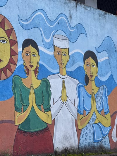 A mural on a rough-textured, light blue concrete wall depicts three figures with yellow faces and dark hair, stylized with simple geometric shapes. The figures are positioned centrally and are all depicted with hands pressed together in a gesture of greeting or prayer. To the left, a female figure wears a green blouse with puffed sleeves and a dark red skirt. Her earrings are yellow and jagged, and her hair is dark and styled in waves. A large, stylized yellow sun with red triangular rays is positioned behind her left shoulder. In the center, a male figure wears a white robe and a white cap with a textured brim. To the right, a female figure wears a bright blue saree with a pattern of concentric circles. Both female figures have a red dot on their forehead. The background behind the figures features stylized blue waves. The mural is located in Dondra, Sri Lanka, a coastal town in the Southern Province known for its lighthouse and ancient Dondra Temple.