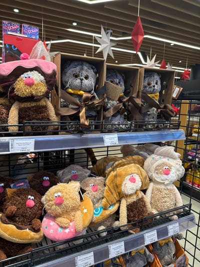 A shelf is filled with stuffed animal toys. Gray stuffed cat toys are displayed in wooden boxes adorned with brown ribbons. Adjacent to the cat toys are stuffed cat toys designed to resemble various foods like donuts and croissants. These are arranged in rows on the shelves. Above the display are more rows of shelves. In the background, decorative stars and diamonds hang from the ceiling.