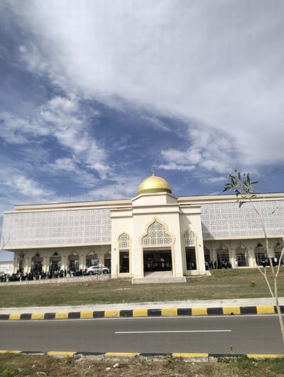 The image captures the exterior of a mosque in Reuleut Timu, Indonesia, under a partly cloudy sky. The mosque features a prominent golden dome and a facade adorned with intricate white geometric patterns. Several people are visible gathered in front of and inside the mosque, some appearing to be entering or exiting. A silver car is parked near the entrance. The mosque is situated along a road with a distinct yellow and black striped curb. The surrounding area includes a grassy lawn and a young tree.