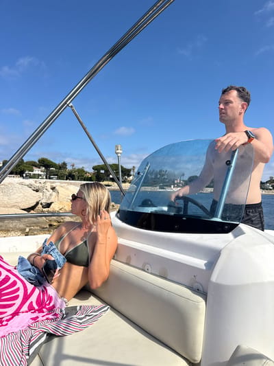 Two individuals are aboard a white motorboat on a sunny day. A male, shirtless and wearing black patterned swim trunks and an orange-faced watch, is at the helm, holding the steering wheel. A female, also shirtless, is seated to his left, wearing a dark green bikini top and sunglasses. She holds a smartphone and a denim jacket. A vibrant pink and white patterned towel and a striped towel are draped over her lap. The boat is on the water, with a coastline featuring rocky formations, trees, and houses visible in the background. The sky is a clear blue with a few wispy clouds. The scene appears to be in Antibes, France, given the distinctive coastline and architecture.