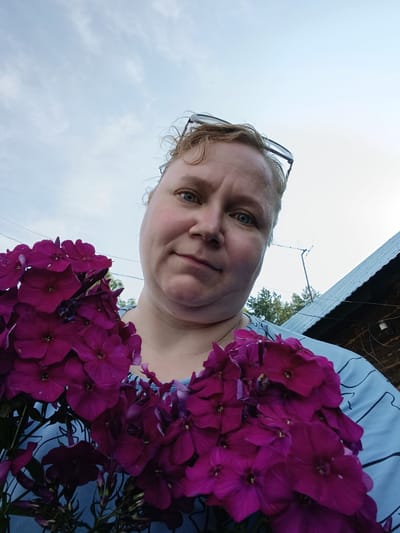 A medium shot shows a woman holding a bouquet of vibrant purple flowers, likely phlox, in front of her. Her head is slightly tilted, and she is looking directly at the camera with a neutral expression. She has blonde, curly hair and is wearing glasses perched on top of her head. Her attire is a light blue shirt with a dark blue pattern.
The background suggests an outdoor, possibly rural, setting. To the right, a dark wooden structure with a corrugated metal roof is visible, hinting at a house or outbuilding. A tall antenna extends upwards into the pale, cloudy sky, typical of a location outside a major city. In the distance, the tops of green trees can be seen. The overall lighting indicates it's daytime, possibly late afternoon or early evening, given the soft light. The scene is consistent with a home or garden environment in a village or smaller town, such as Зипуново, Russia.