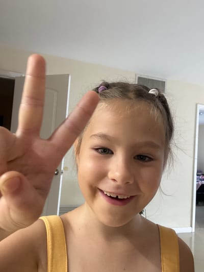 This media file is a selfie of a young girl with blonde hair styled in pigtails. She is smiling broadly, showing her teeth, and making a peace sign with her right hand, which is positioned in the foreground. Her eyes are blue and she has a playful expression. She is wearing a yellow top. The background appears to be indoors, with a white wall and a door visible. A light fixture is also discernible on the ceiling. The lighting suggests it is daytime. The girl is likely at home, given the indoor setting. The overall mood of the image is cheerful and lighthearted. There is no visible text.