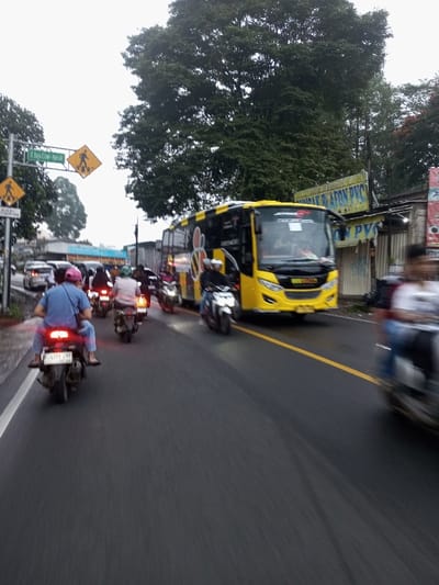 The image displays a traffic scene on a wet asphalt road in Dawathang_Dorjibi_ Kashingtsawa, Bhutan, under an overcast sky. The road features a solid yellow center line and a white shoulder line on the left.

Multiple vehicles are present. In the foreground and mid-ground, numerous motorcycles are navigating the roadway. The closest visible motorcycle, a dark-colored scooter, has a rider in a blue shirt and a pink cap, and a passenger, both wearing sandals. The scooter's taillight is illuminated, and its rear license plate reads "F 5391 KP". Other motorcycle riders are visible behind, some wearing helmets and one in a white shirt.

A large, modern yellow bus with black accents and "JAVANEST" branding is prominent on the right side of the road, facing the viewer's left. It appears to be in an adjacent lane or part of slow-moving traffic. Its front is distinctly visible with large headlights. Further down the road, additional cars are visible in the distance on the left.

Roadside features include a green street sign with white text reading "Jl. Raya Ciapus - Parakan" on the left, mounted below a yellow diamond-shaped pedestrian crossing sign. Another identical pedestrian sign is visible further along the road. The upper background is dominated by dense green foliage from large trees. On the right, behind the bus, a commercial structure with a corrugated metal facade displays a sign advertising "JUAL PLAFON PVC". Traffic appears to be moving slowly.
