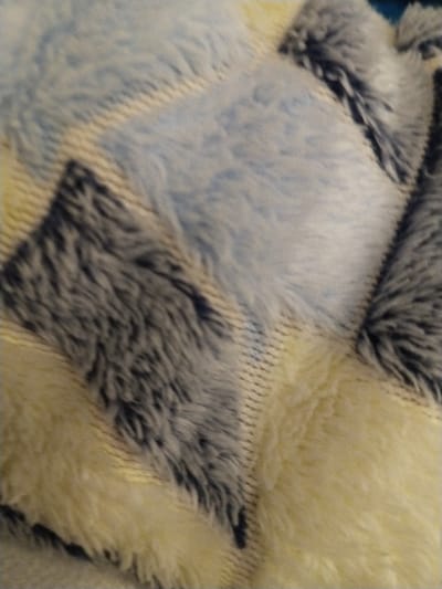 A close-up shot reveals a plush, textured surface with a geometric pattern. Diamond-shaped patches in light blue, creamy white, and gray-blue hues create the design. Each patch features a long, fuzzy pile, giving the impression of softness and depth. Visible stitching or seams delineate the patches. The fabric appears densely woven and luxurious.