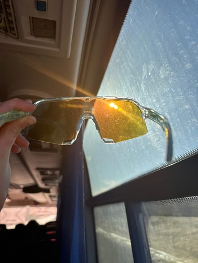 A pair of yellow-tinted sunglasses is held up to a car windshield. The sun is reflecting brightly off the lenses of the glasses. The interior of the car is visible, including the dashboard and the sun visor. The location, Челва, Russia, cannot be determined from this image.