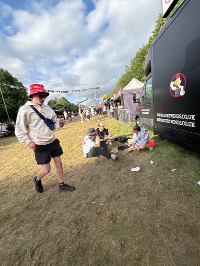 An outdoor event showcases attendees and food vendors. A man in a "TWAT" bucket hat and casual wear stands prominently. Nearby, people sit on the grass, some near a Gurt Wings food truck. Festive bunting adds to the atmosphere, and other vendors are visible in the background. The sky is partly cloudy, suggesting a pleasant day for the event.