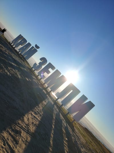 A wide-angle, low-angle shot captures a large, white, three-dimensional Cyrillic inscription that reads "КРАЙ ЗЕМЛИ" (Krai Zemli), meaning "Edge of the Earth." The letters are positioned on a sandy, uneven surface that slopes downwards towards a body of water, likely the Arctic Ocean, visible in the background. The sun, a bright glare in the upper center of the frame, casts long, sharp shadows of the letters across the sand, indicating it is likely morning or late afternoon. A single person, silhouetted against the bright sky, stands on the left side of the frame, appearing to take a photograph of the inscription. The environment is a coastal area with sparse vegetation along the water's edge. The sky is clear and blue, with a subtle gradient towards a lighter hue near the horizon. The image is rotated counter-clockwise.