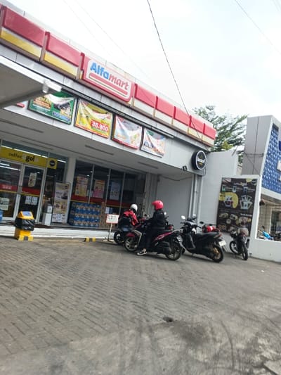An exterior view depicts the entrance to an Alfamart convenience store in Margahayu, Indonesia, under an overcast sky. The store features a red and white striped facade with the "Alfamart" logo prominently displayed. Below the logo, multiple promotional banners are affixed. To the left of the main entrance, a "coffee go!" sign is visible. The storefront consists of large glass panels revealing interior shelving with various products and stacked cartons, possibly containing bottled water. A yellow and black garbage bin is situated to the left of the entrance.
The forecourt is paved with gray interlocking bricks, showing some dark stains. Four motor scooters are parked in front of the store. Two individuals are seated on separate scooters; one wears a red helmet and black jacket, while the other wears a white helmet and red jacket, with a red delivery bag on the back of their scooter. A third scooter is parked to their immediate right, and a fourth is partially visible further right. A small white sign with Indonesian text stands near the scooters. To the right of the Alfamart, a black circular sign for "beans! coffee & eateries" is mounted on the building wall. Further right, a portion of another building with a blue textured facade is visible, alongside green tree foliage. Overhead, multiple thin power lines traverse the frame.