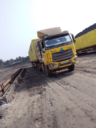 The image depicts an outdoor industrial or mining scene in Hoche, Nigeria, under an overcast or hazy sky. The primary subject is a yellow Sinotruk Howo heavy-duty truck, positioned at an angle on an uneven, dark dirt road with visible tire tracks. The truck features a raised, yellow, high-sided cargo body or trailer, typical for bulk material transport. Its front bumper displays minor deformation, and a Nigerian license plate, "04A12EB," is affixed below the grille.

To the left of the truck, a long, brown, rusty metal structure, resembling a weighbridge or a component of a loading facility, is partially visible, supported by concrete blocks embedded in the dirt. In the background on the right, another yellow truck or trailer, also with a high-sided cargo body, is parked, seemingly laden with a dark, granular material, likely coal or ore, which is also piled in large mounds further behind. Distant palm trees or similar tropical vegetation are visible in the far background on the left horizon. No individuals are discernible in the frame.