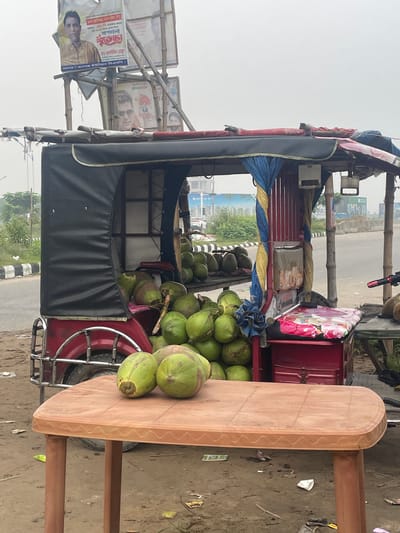 Here's a concise description of the image:
The image shows a coconut vendor's rickshaw in what appears to be Purbachal, Bangladesh. The rickshaw is loaded with fresh green coconuts. A makeshift table is attached to the rickshaw, holding additional coconuts. The rickshaw has a black weather shield. Political posters are attached to a bamboo structure on the rickshaw. The environment suggests an urban or suburban setting with a road.