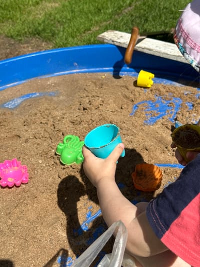 This media file shows a close-up of a child's hands playing in a sandbox. The child is wearing a pink and navy striped shirt and their skin is fair. The sandbox is blue and filled with sand. Several sand toys are visible, including a yellow castle mold, an orange shell-shaped mold, a green octopus mold, and a pink flower-shaped mold. A small wooden shovel is also present. The child is holding a turquoise cup and appears to be scooping sand. The scene is outdoors, with green grass visible to the left of the sandbox, suggesting a sunny day. The lighting indicates it is daytime, likely late morning or early afternoon, with strong shadows cast on the sand. The overall impression is of a child engaged in playful activity in a backyard or park setting. No visible text is present in the image.