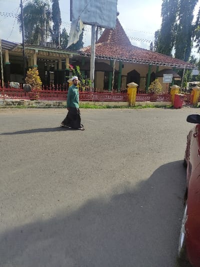 The image, taken in Situbondo, Indonesia, captures an outdoor scene under bright daylight. In the foreground, an individual stands on an asphalt road, looking towards the camera with a neutral expression. The person, appearing to be a man, wears a light green long-sleeved shirt, dark pants, and a white cap, possibly a peci. His hands are clasped loosely in front of him.

To the left of the road, a long, single-story building with a prominent reddish-brown tiled roof extends across the midground. The building features light-colored walls and supports, with several open archways along its facade. A decorative red metal fence or railing runs along the front of the building, separating it from the road. Positioned beside this fence are several yellow and red waste bins. Behind the building and further in the distance, numerous tall green trees are visible, suggesting a lush environment. A large, square signpost or billboard structure stands on the left side of the road, partially obscuring the view of the building.

The road surface is dark asphalt, and distinct, elongated shadows, cast by the person, the building, and an unseen object to the right, indicate strong sunlight, suggesting it is either mid-morning or late afternoon. A portion of a red car is visible in the upper right corner of the frame, parked or stopped near the road's edge. The sky above appears clear and bright. While there are signs on both the building and the large signpost, any text on them is unreadable due to the distance and resolution.