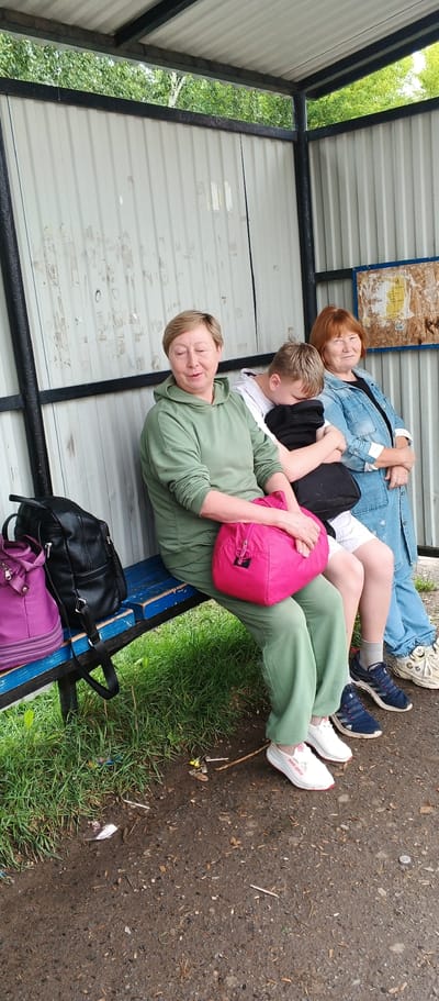 Three individuals are seated on a blue wooden bench under a metal and corrugated metal shelter. The shelter has a white corrugated metal wall behind the bench and a white corrugated metal roof supported by black metal posts. Green foliage is visible above the shelter.
On the left, a woman with short, light brown hair wearing a green hooded sweatshirt and green sweatpants is seated. She holds a bright pink duffel bag on her lap. To her left, on the bench, are a black leather backpack and a purple shoulder bag.
In the center, a young person, likely a boy, with light brown hair is leaning forward, resting their head on a black backpack on their lap. They are wearing a white t-shirt and white shorts.
On the right, a woman with short, reddish-brown hair wears a blue denim jacket over a dark shirt and blue jeans. She is looking towards the right of the frame.
The ground in front of the bench is a mixture of dark, wet soil and gravel, with patches of green grass to the left and some litter visible on the ground. The overall scene appears to be a bus stop or waiting area.