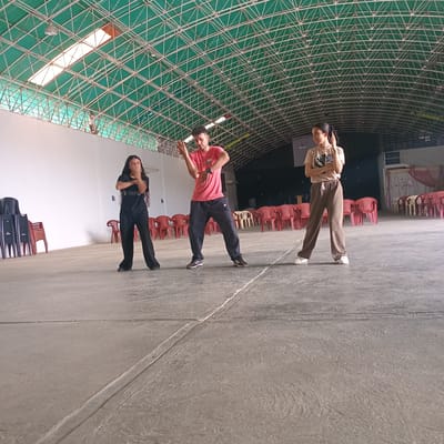 The image depicts three individuals in what appears to be a large indoor space, possibly a gymnasium or warehouse. The location is identified as Duaca, Venezuela. The space has a high ceiling with a complex lattice structure, partially illuminated by rectangular light fixtures. The floor is concrete, marked by linear divisions. Rows of red plastic chairs are visible along the wall, with some stacks of black chairs. The three individuals are positioned in a line across the frame. The person closest to the viewer is wearing black clothing and has long hair. The person in the middle wears a red shirt and dark pants. The person furthest away is wearing a tan shirt and brown pants. All three appear to be posing for the photograph. Their actions do not suggest any specific interaction beyond the photo.