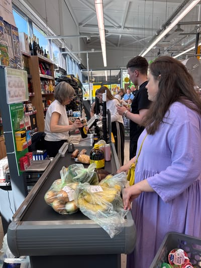 The image, rotated 90 degrees clockwise, depicts a busy grocery store checkout lane in Finskiy Briz, Russia, likely a Lenta supermarket.
In the foreground, a woman with long, dark hair, wearing a lavender dress and carrying a yellow shoulder bag, is placing groceries onto the dark grey conveyor belt. Her items include plastic bags containing bananas and onions, a bottle of kvass, and bread. A grey shopping basket with a "VIOLA Very berry" yogurt container is partially visible next to her.
Behind her, a man with short, dark hair and a black t-shirt waits in line. Further down the line, a cashier, a woman with short, light-colored hair and glasses wearing a white t-shirt, is actively scanning items. She holds a bottle similar to the one on the conveyor belt, and more bread products are visible next to the scanner.
Adjacent to the checkout, display stands offer impulse buys, including SIM cards, indicated by "ПЕР СИМ-КАРТЫ" text. Overhead, bright fluorescent lights illuminate the store's interior, revealing exposed ceiling structures and ductwork. Large windows allow natural light in.
Other visible text includes "KBAC" on a bottle, "лента" (Lenta) on a sign, and fragmented texts such as "23:00", "Вого, вот оно", "1-3", "Манай", and "БЕЗ ПОСЕ" on various product labels or store signage. The scene captures a typical daytime shopping experience, with customers engaged in the transaction process.