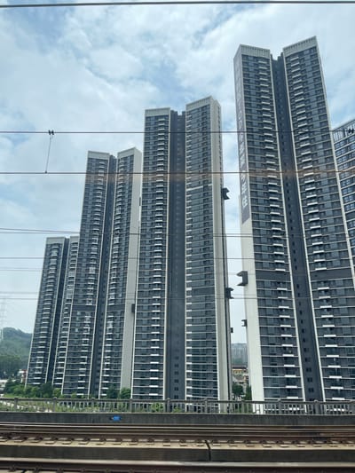 In Dongguan, China, a series of identical, modern high-rise apartment buildings dominate the skyline under a cloudy sky. The buildings are tall and slender, with dark gray facades punctuated by rows of windows and white vertical accents. Some of the buildings display large banners with Chinese text and a logo that reads "TOD". In the foreground, railroad tracks run horizontally across the image, suggesting the perspective is from a moving train. Overhead, power lines stretch across the frame, partially obscuring the buildings. The landscape behind the buildings includes rolling green hills and some scattered trees. A concrete barrier with a metal railing separates the railroad tracks from the area in front of the buildings.