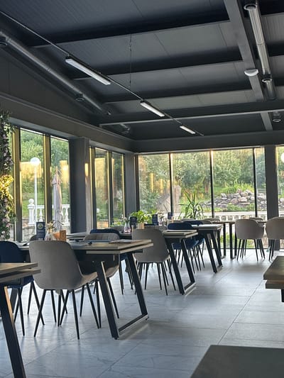 The image depicts the interior of a modern restaurant or cafe with large windows overlooking a lush, green outdoor area. The restaurant features contemporary furniture, including sleek tables with dark metal legs and comfortable upholstered chairs in shades of gray and blue. Long, rectangular fluorescent lights are suspended from the dark, industrial-style ceiling, which also has exposed ventilation pipes.
The floor is tiled with large, light gray rectangular tiles. On the tables, there are place settings with cutlery and what appear to be menus. Decorative elements include small potted plants and floral arrangements, one of which, a vertical cascade of pink flowers, is visible on the left side of the image.
Through the expansive windows, a sunlit garden or park is visible, with trees, stones, and a white balustrade. A closed umbrella and a lamppost are also noticeable in the outdoor scene. The lighting suggests it is either late afternoon or early morning, with a warm, golden hue illuminating the outdoor landscape. There are no people visible in the image. The overall impression is one of a sophisticated and tranquil dining environment with a strong connection to nature through its extensive window views.